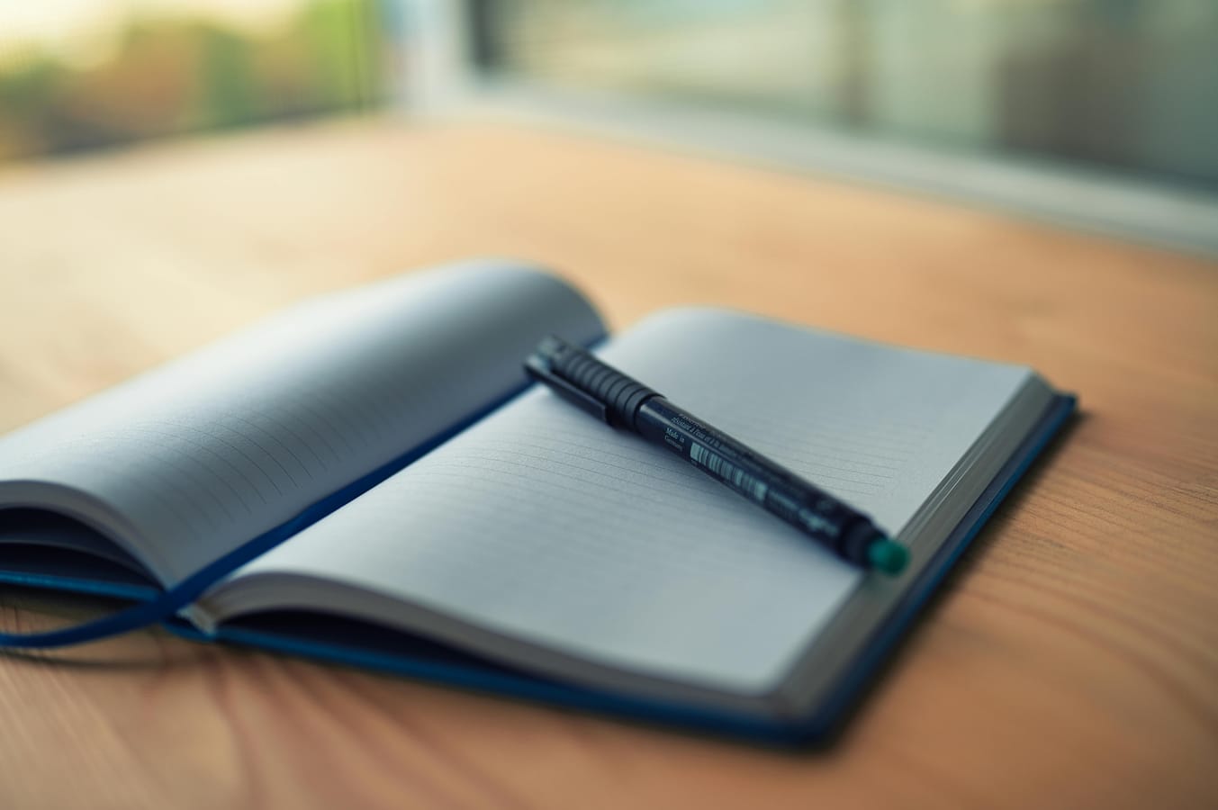 An open notebook with a pen and a glass of water on a wooden desk in soft morning light.