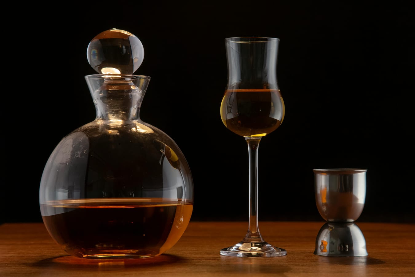 A pint glass, wine glass, and shot glass arranged on a wooden bar with measuring jiggers beside them.