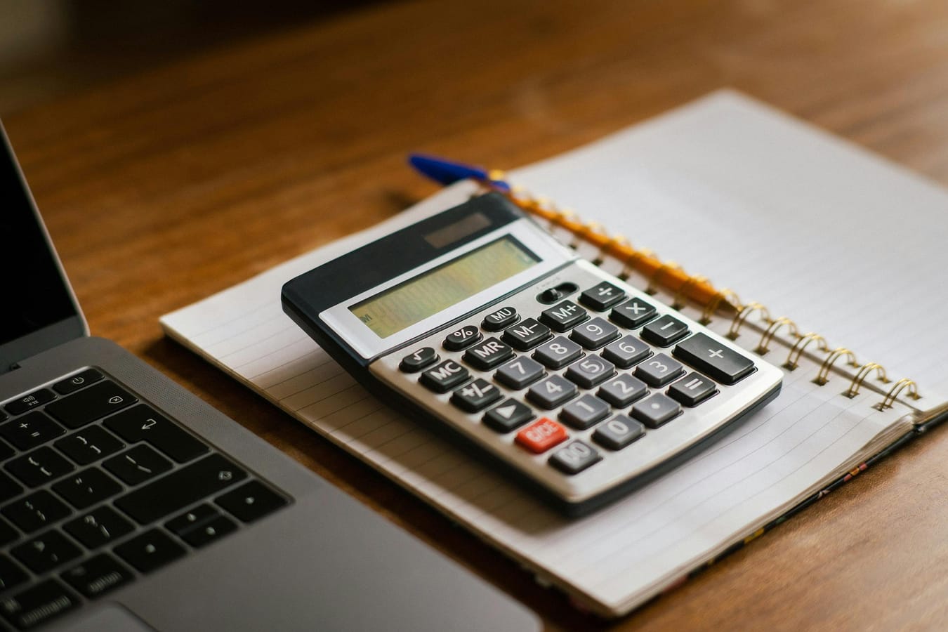 A calculator and a pint glass on a wooden table beside a notepad with handwritten calculations.