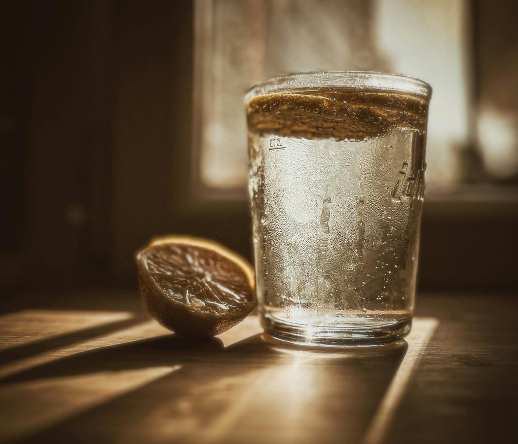 A glass of clear electrolyte drink and a sliced lemon on a wooden table in morning light.