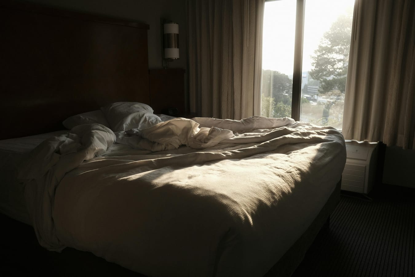 An unmade bed in pre-dawn darkness with a window showing the first hint of morning light.