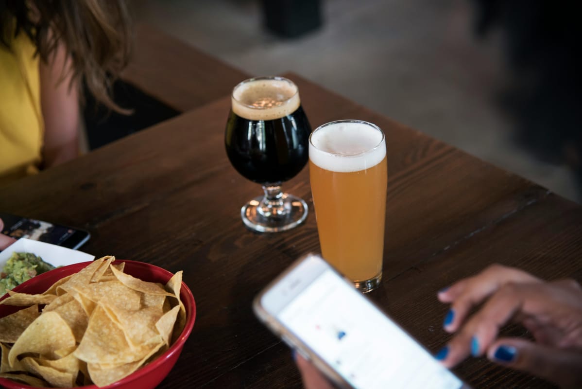 A smartphone on a wooden table beside a glass of beer.