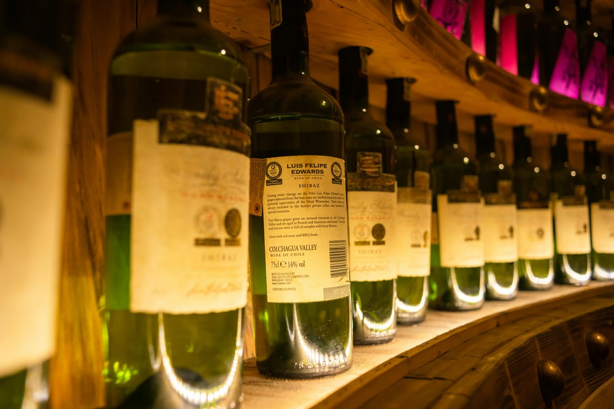 A row of wine bottles on a wooden shelf, ranging from light to dark.