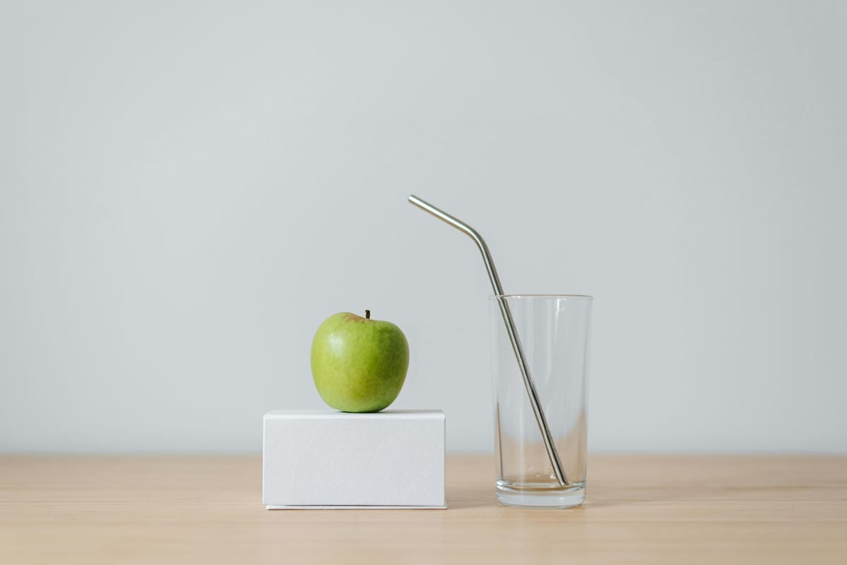 An apple beside a glass of water on a wooden table in natural light.
