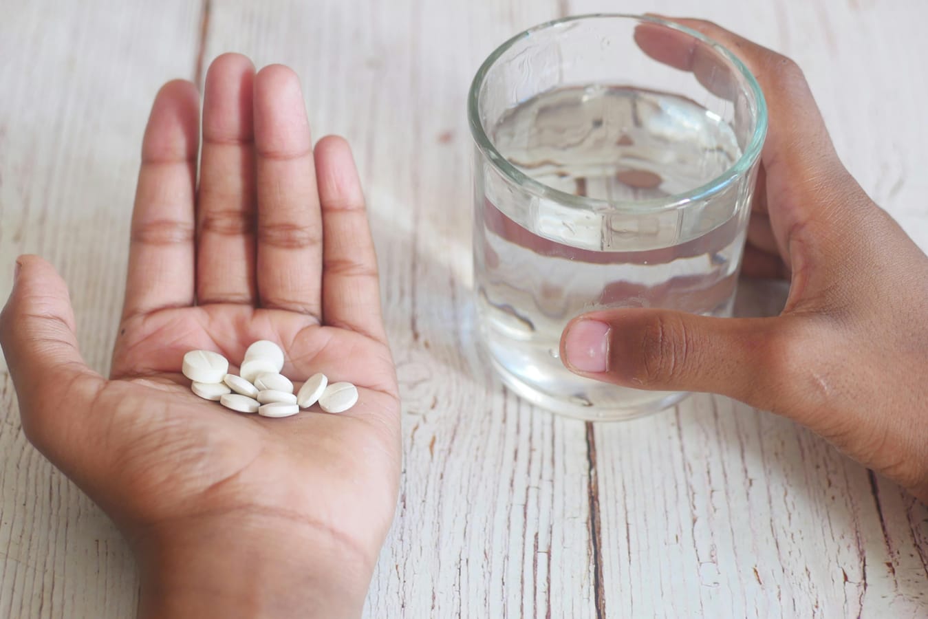 A single white tablet beside a glass of water on a wooden surface in soft natural light.