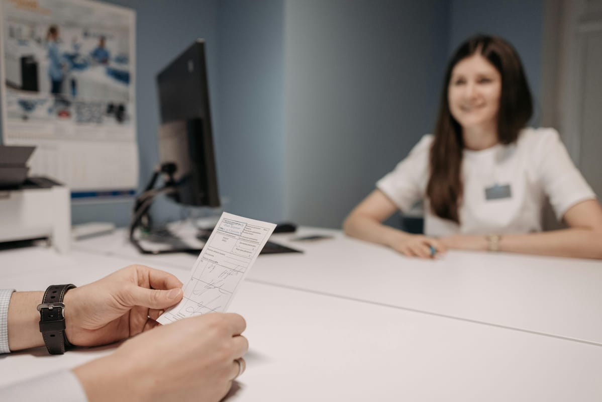 A medical consultation scene with hands on a desk and paperwork, no faces visible.